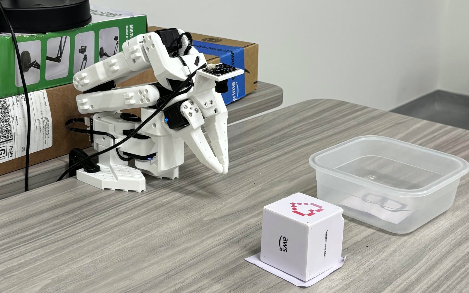 The SO-101 arm on the lab desk beside the marked cube it picks up and the clear container it places the cube into.